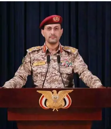 "يحيى سريع، المتحدث باسم الحوثيين، يقف خلف منصة تحمل شعار الجماعة، مرتدياً الزي العسكري وقلنسوة حمراء، خلال إلقاء بيان رسمي."
