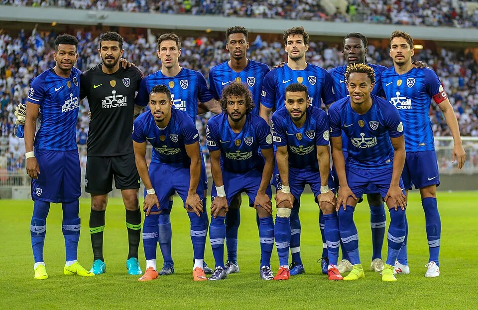 "فريق الهلال يصطف على أرض الملعب قبل مباراة في الدوري السعودي، يرتدون الزي الأزرق مع شعار النادي، وسط حضور جماهيري كبير."